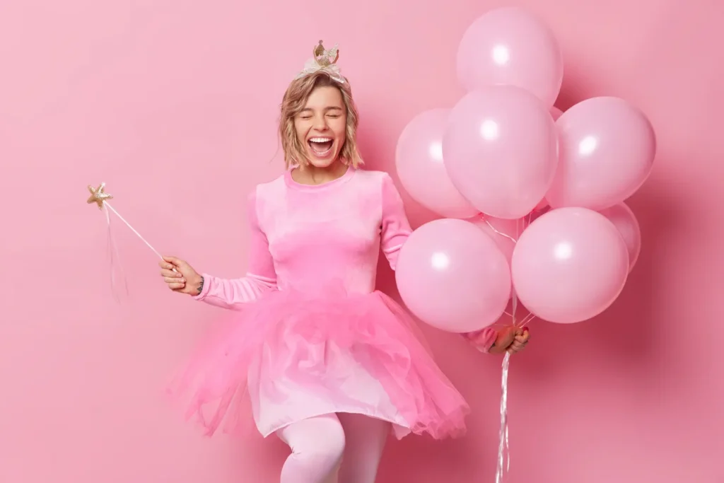 Jovem feliz e sincera com penteado exclama de felicidade, usa coroa e o vestido segura magia e um monte de balões inflados aproveita a celebração e a festa isolada sobre fundo rosa.